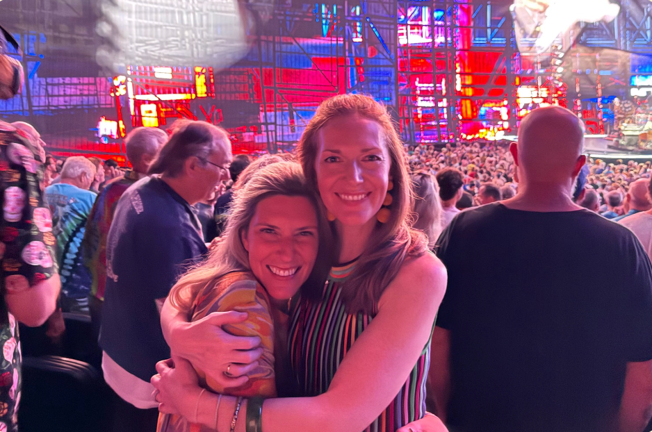 Two friends hug and smile under colorful stage lights at a live music event, both dressed in fun, expressive outfits.