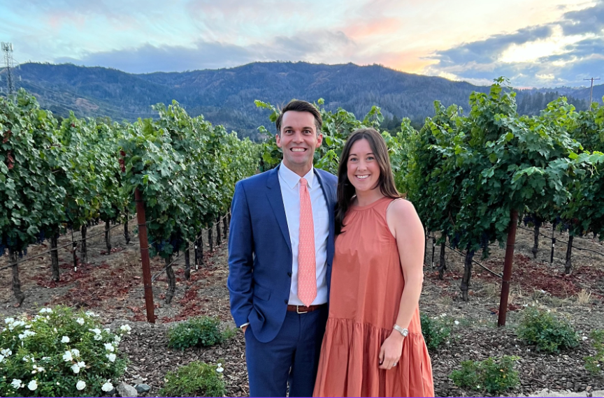 A husband and wife stand together in a scenic wine country vineyard, smiling and relaxed.