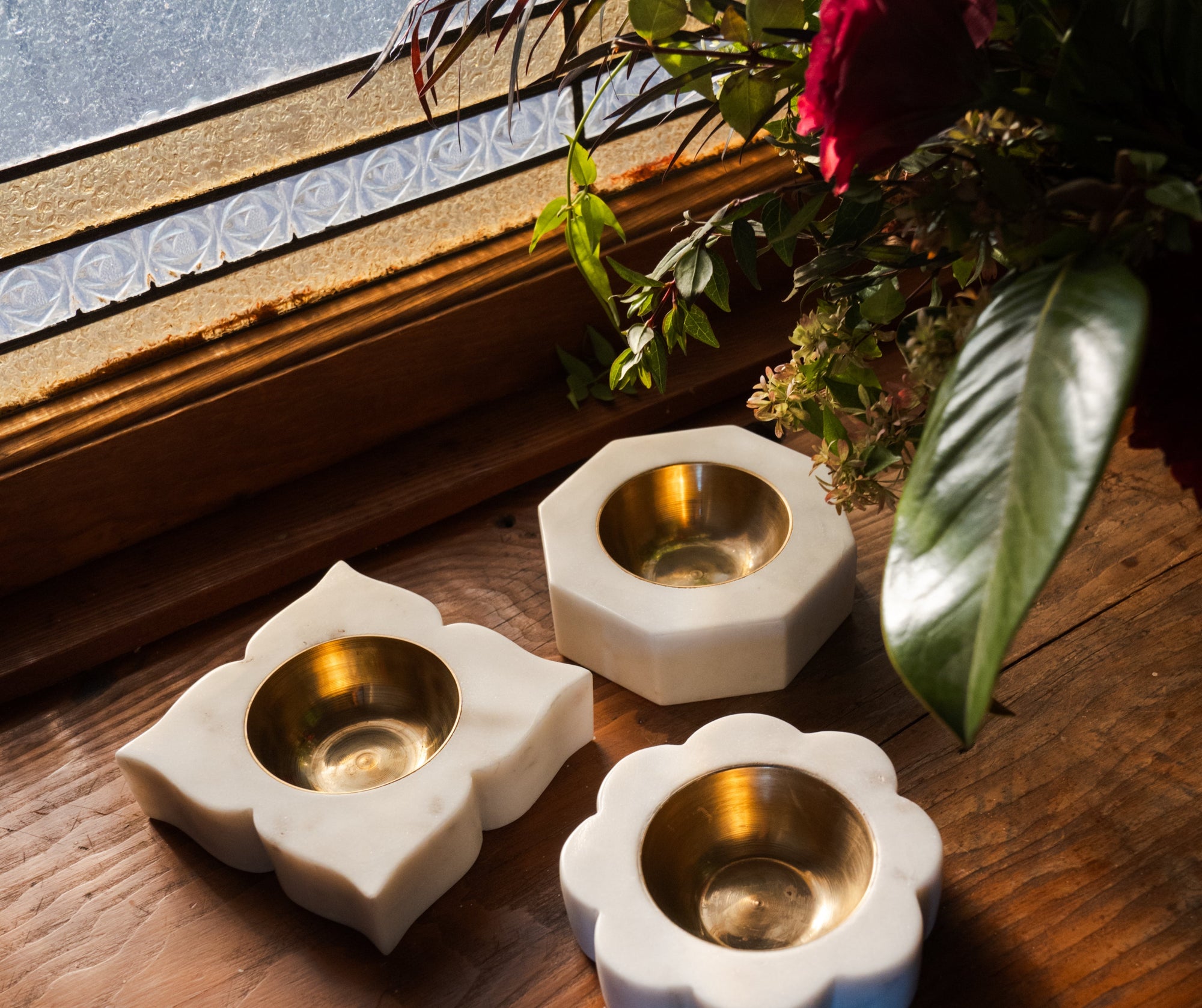 Three marble candle holders with gold interiors on a wooden surface near a window with floral decorations.